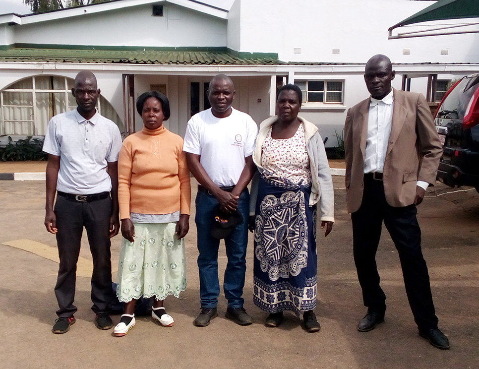 The Executive Director of the KEYS Organization together with four other members are standing on a parking lot in front of a white wooden building with green roof tiles.